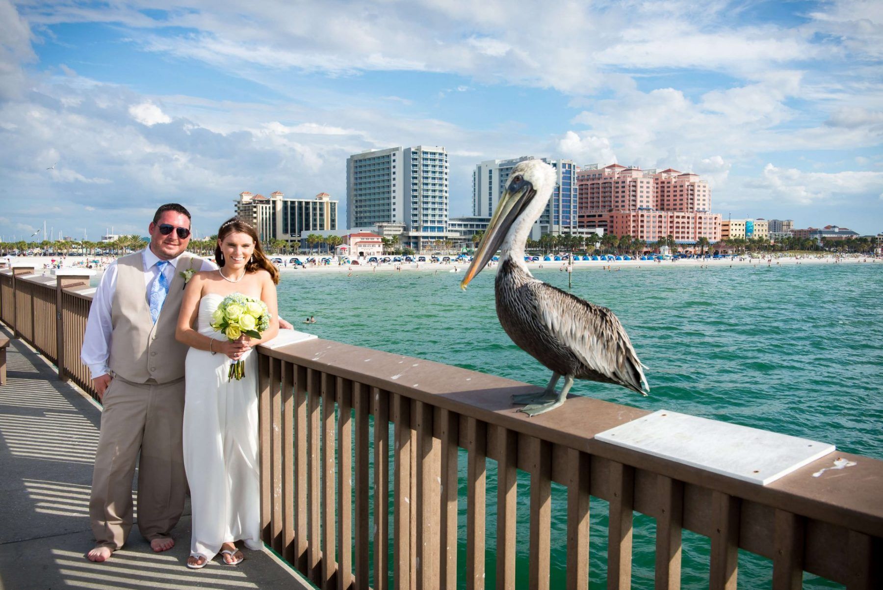 Newlyweds pose on a pier with a pelican, turquoise water, and cityscape background.