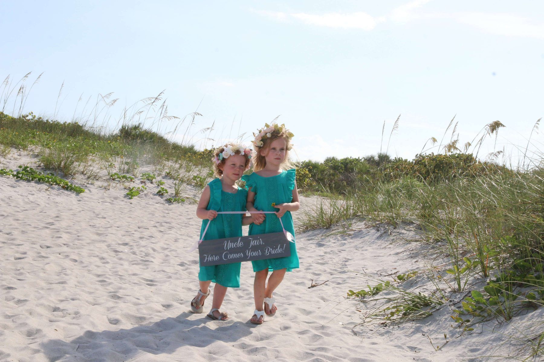 Two girls in teal dresses walk on a sandy path, holding a sign near a beach.