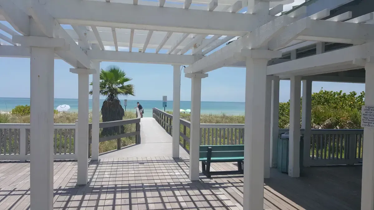 White pergola frames boardwalk leading to the beach. Blue sky and ocean in the distance.