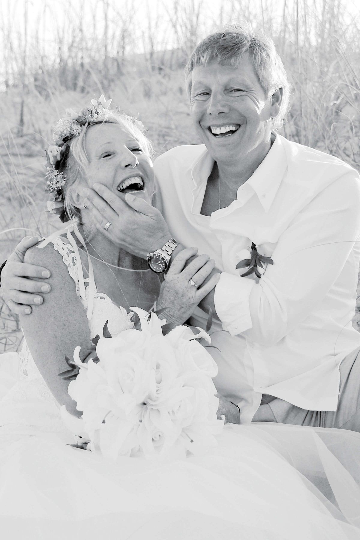 Couple laughing joyfully on a beach. Woman in a white dress holds bouquet, man’s hand on her face.