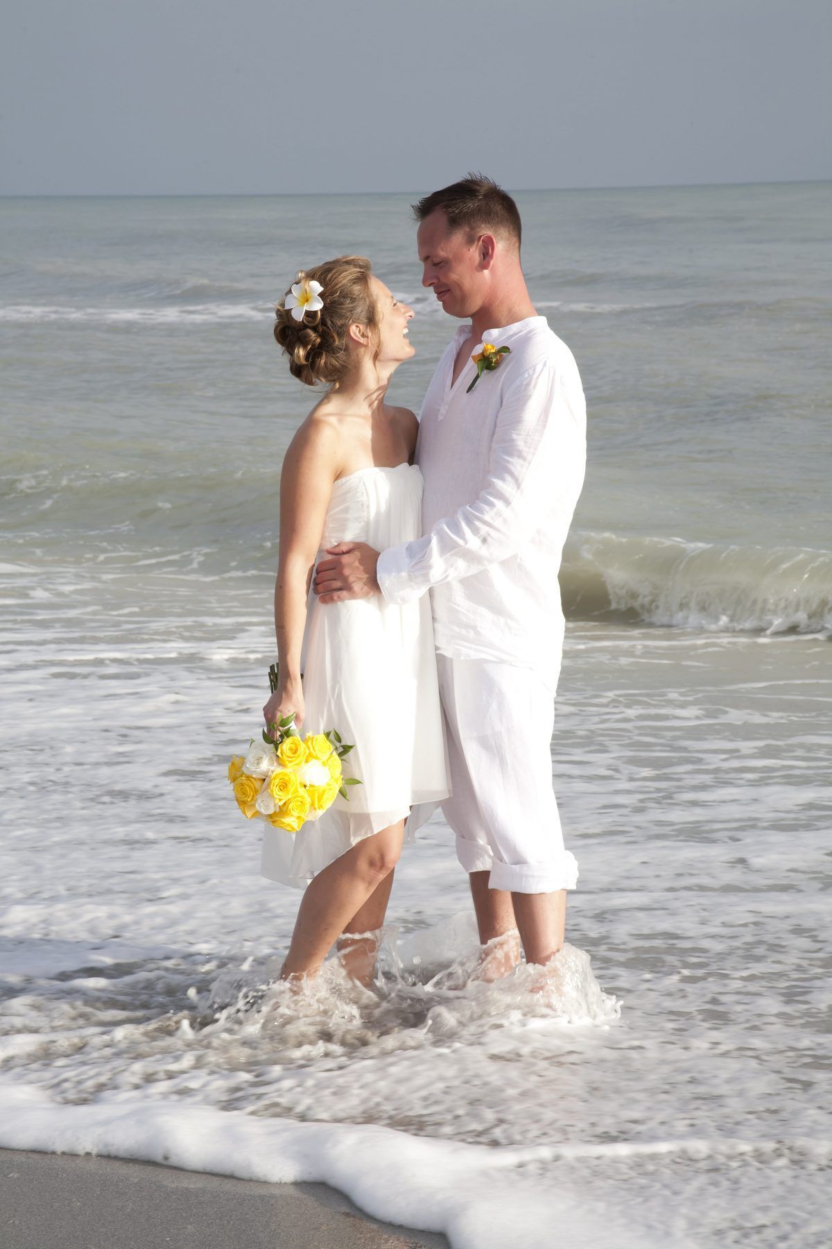 Couple embracing in shallow ocean water at the beach; bride in white dress, groom in white shirt and pants.