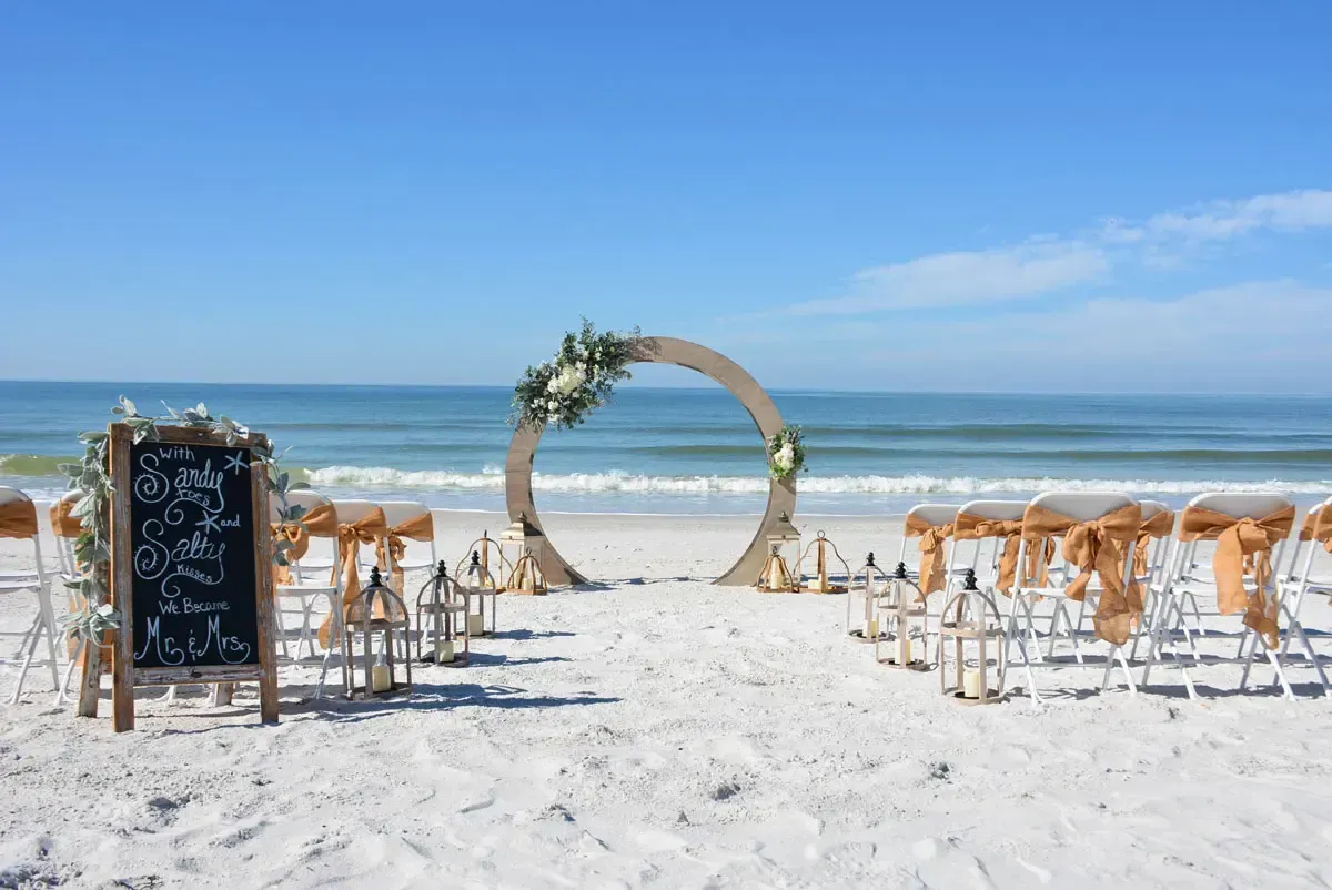 Beach wedding setup with a circular arch decorated with flowers, chairs, and lanterns.