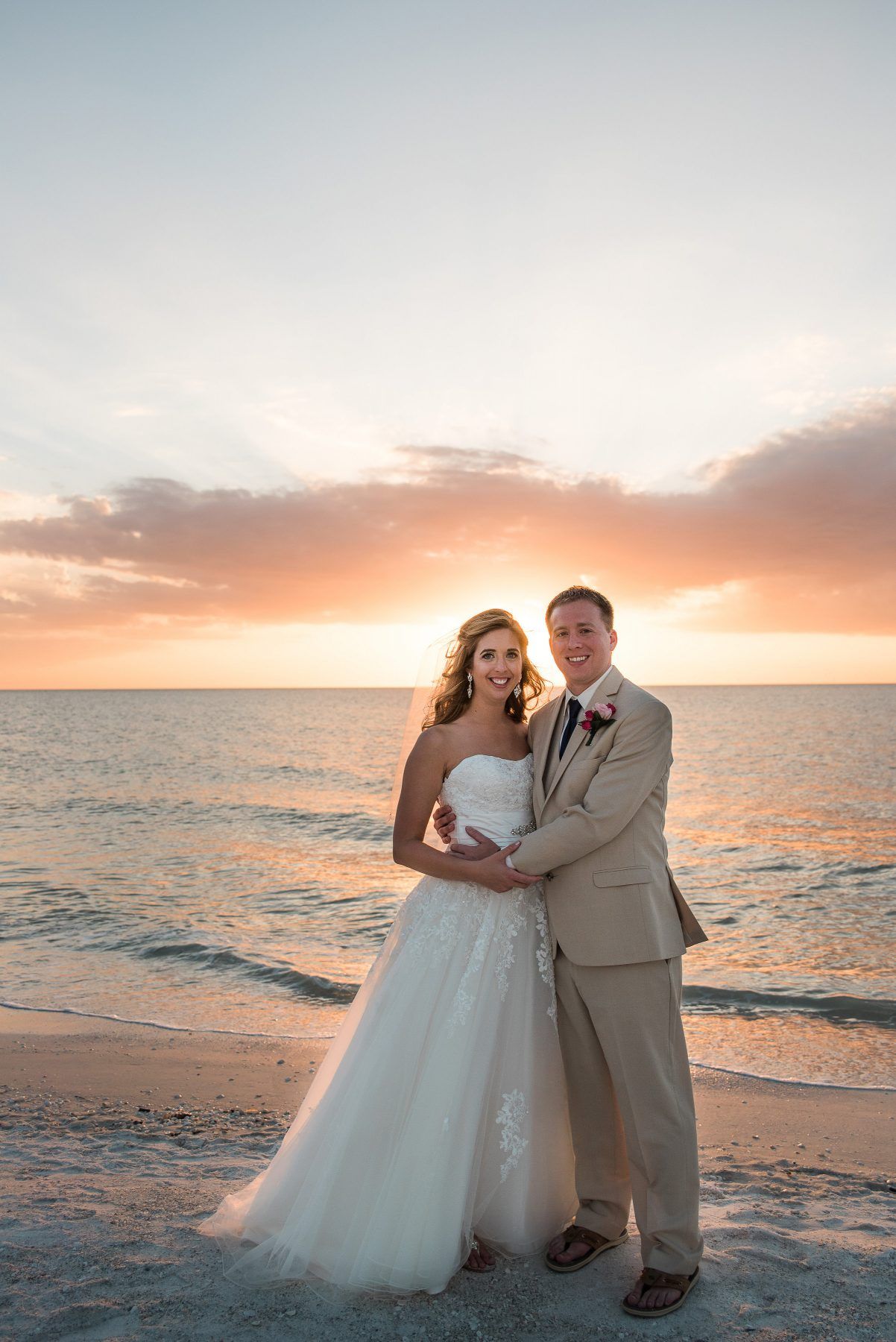 Bride and groom on a beach at sunset, embracing. Dress and suit in warm tones against ocean and sky.