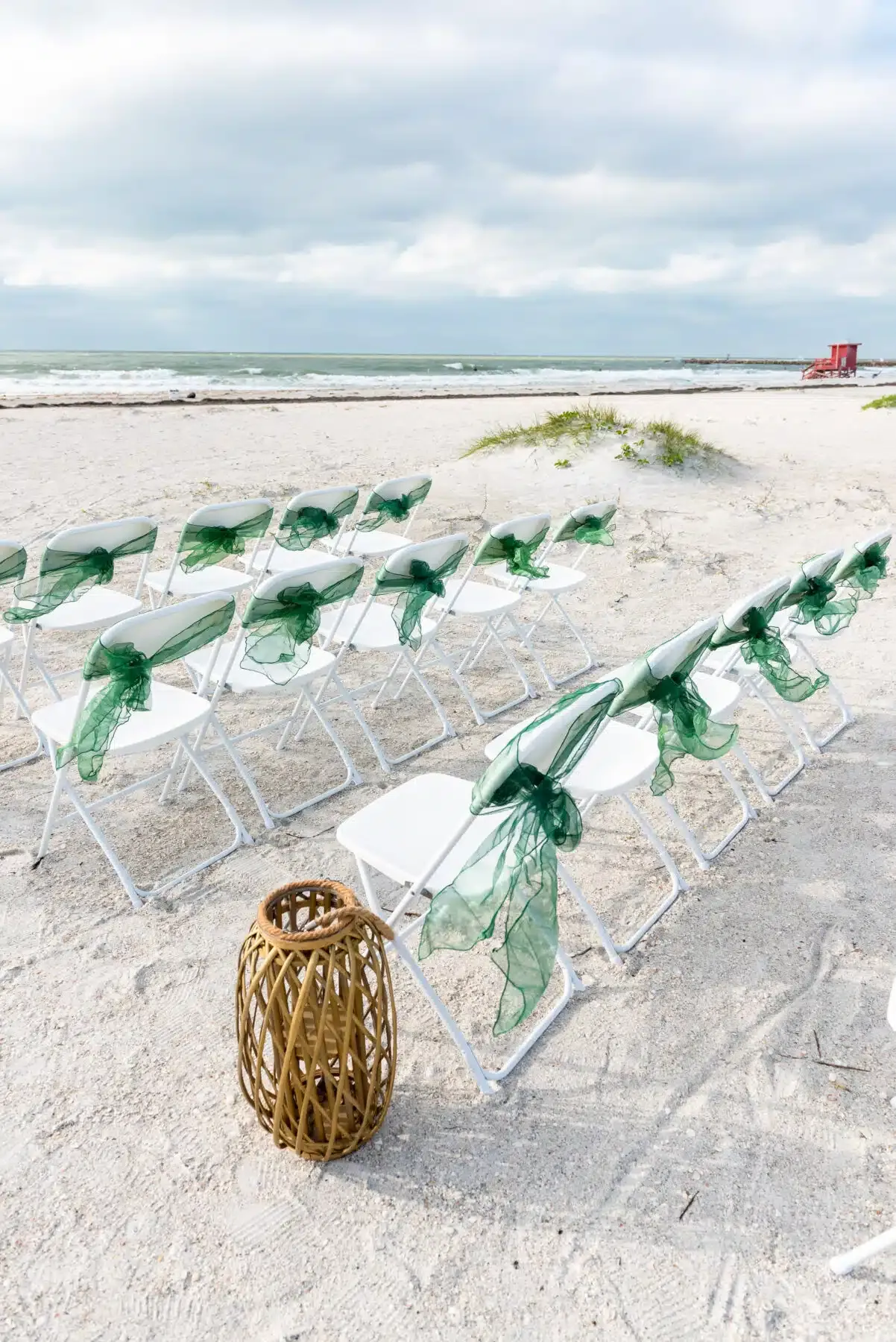 White chairs with green draped fabric on a beach, waves in background, cloudy sky.