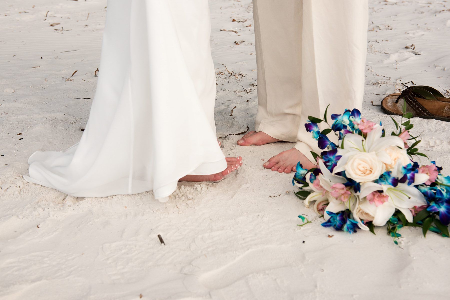 Bride and groom's bare feet on sandy beach next to wedding bouquet; white, blue, and pink flowers.