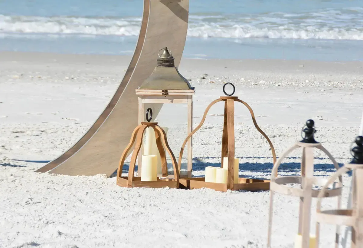 Wooden lanterns with candles on a sandy beach, against a large arch and ocean background.
