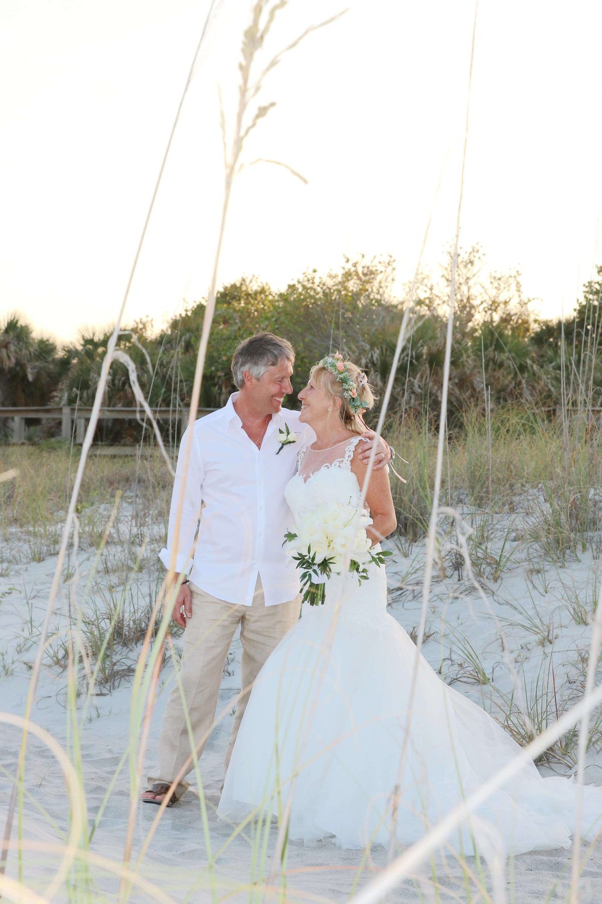 Bride and groom embrace on a sandy beach at sunset, framed by tall grass.