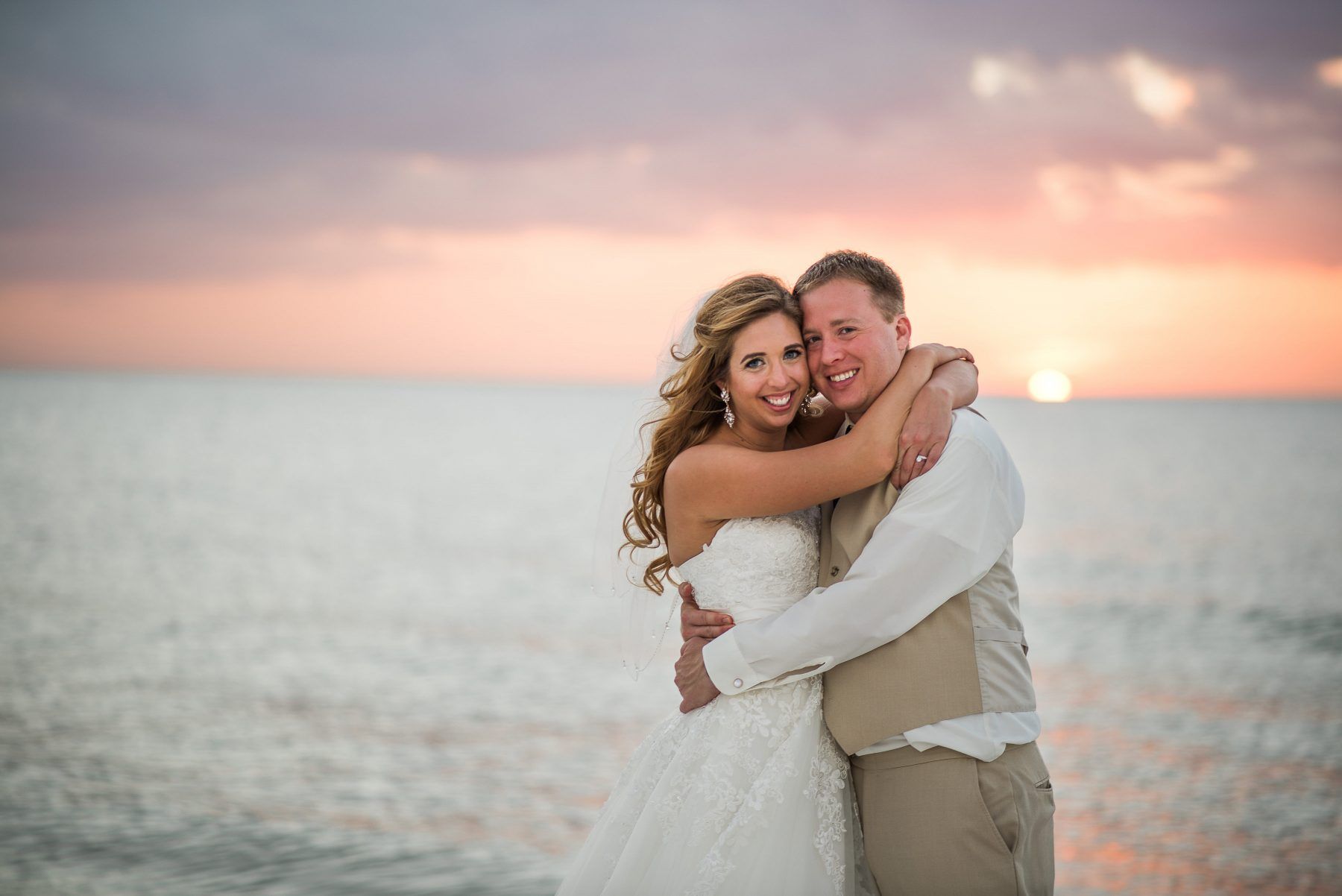 Newlyweds embrace on a beach at sunset. Bride in white gown smiles, groom in vest hugs her.