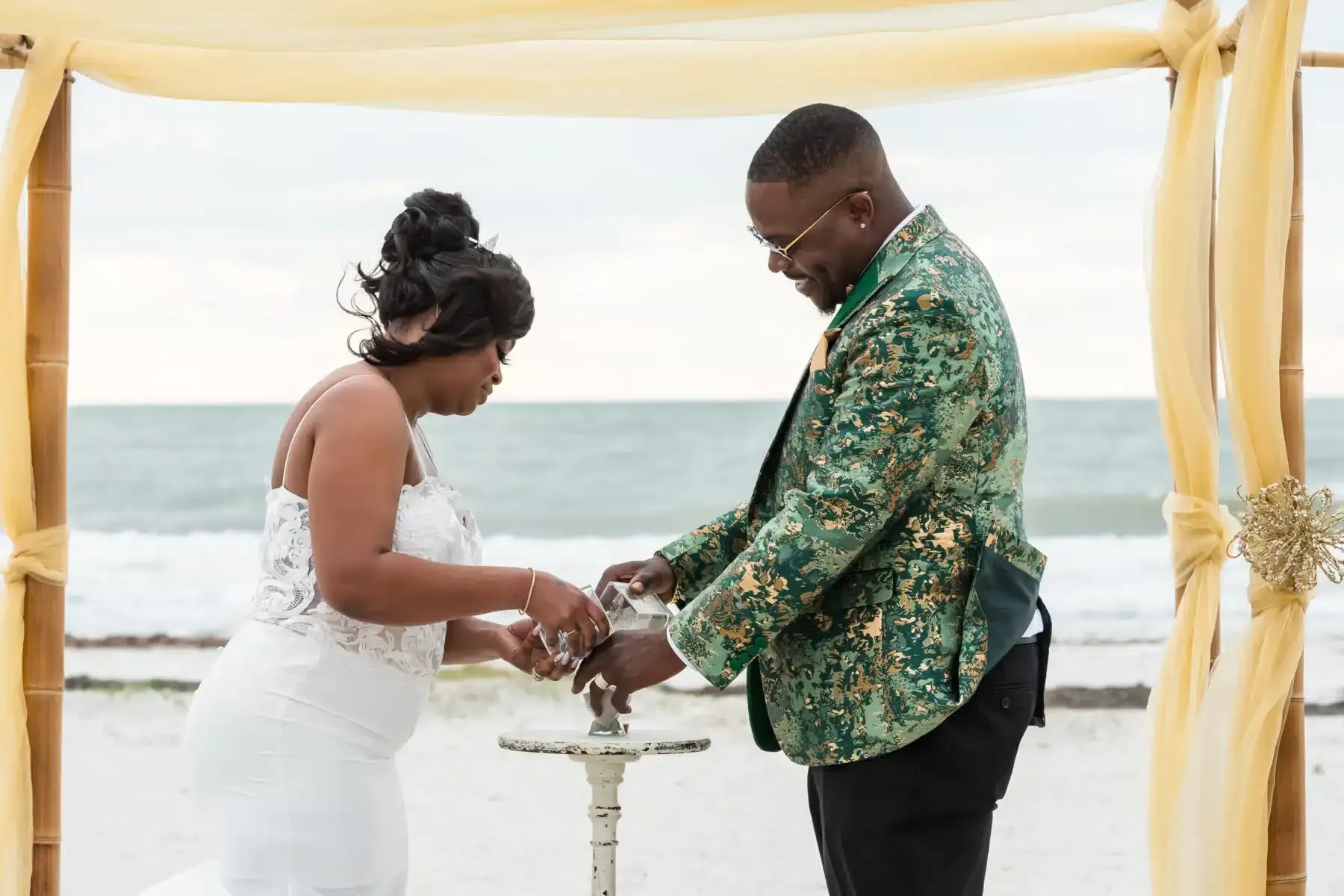 Couple pouring sand during a beach wedding ceremony, under a draped bamboo arch.
