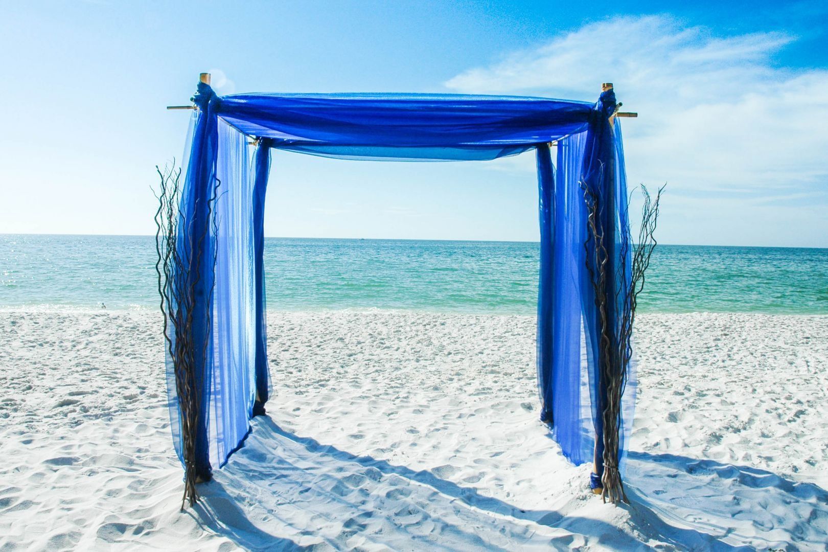 Blue draped wedding arch on a white sand beach, ocean and blue sky in the background.