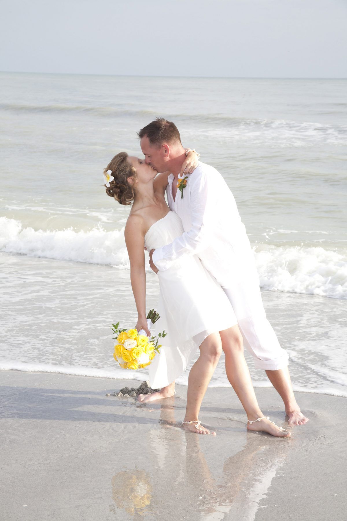 Couple kissing on a beach. Bride in white dress and groom in white shirt and pants, near the water.