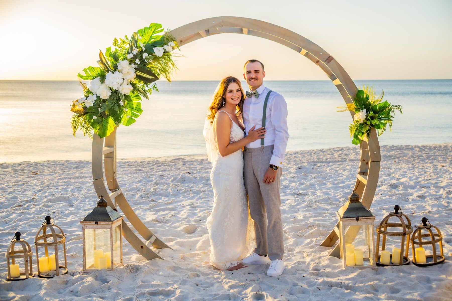 Couple posing under floral wedding arch on beach at sunset.