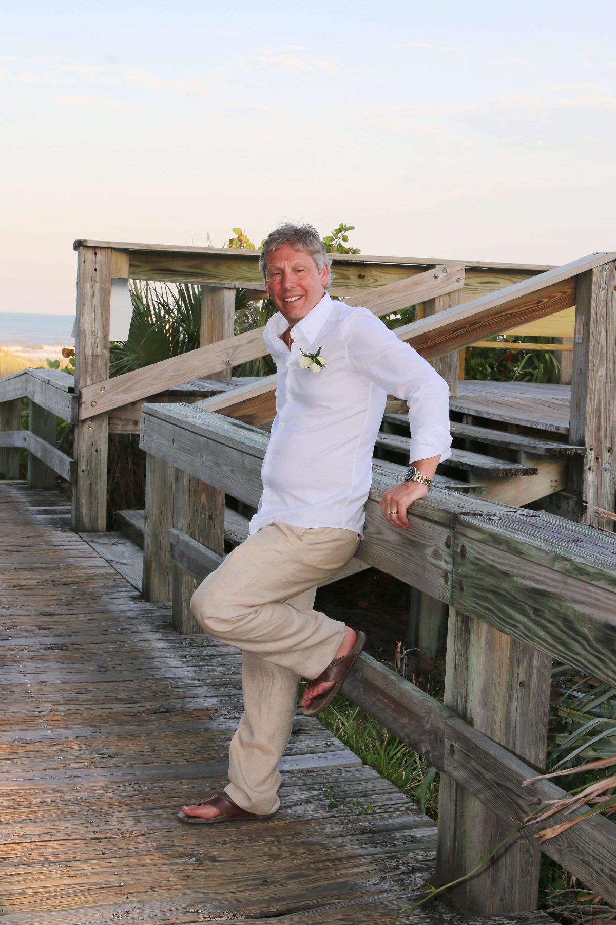 Man in white shirt and khaki pants leans on a wooden railing on a boardwalk; beach in background.