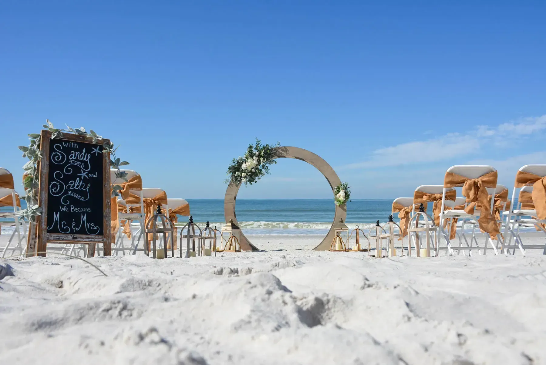 Beach wedding ceremony setup with white sand, blue sky, and ocean in background.