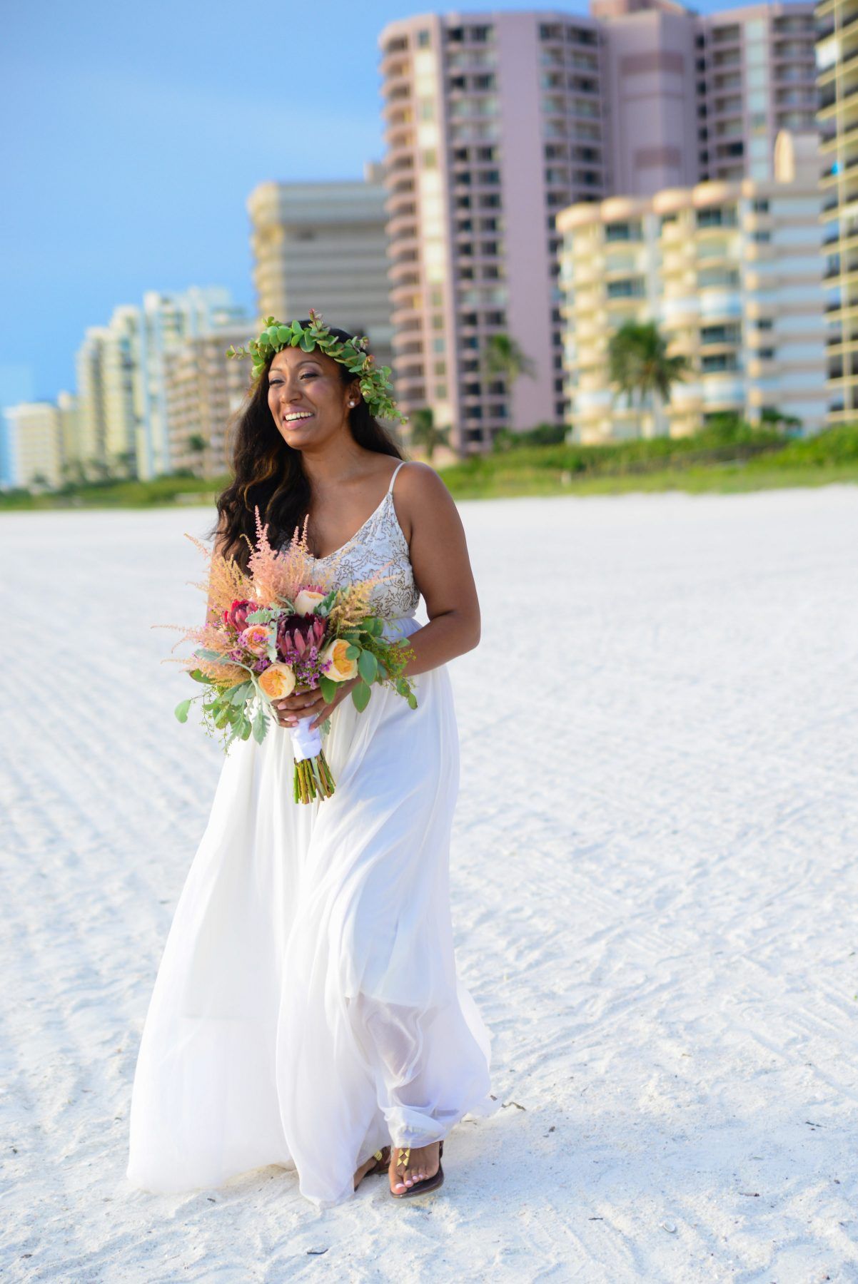 Woman in white dress with flower crown, holding bouquet on a beach, smiling with buildings in background.