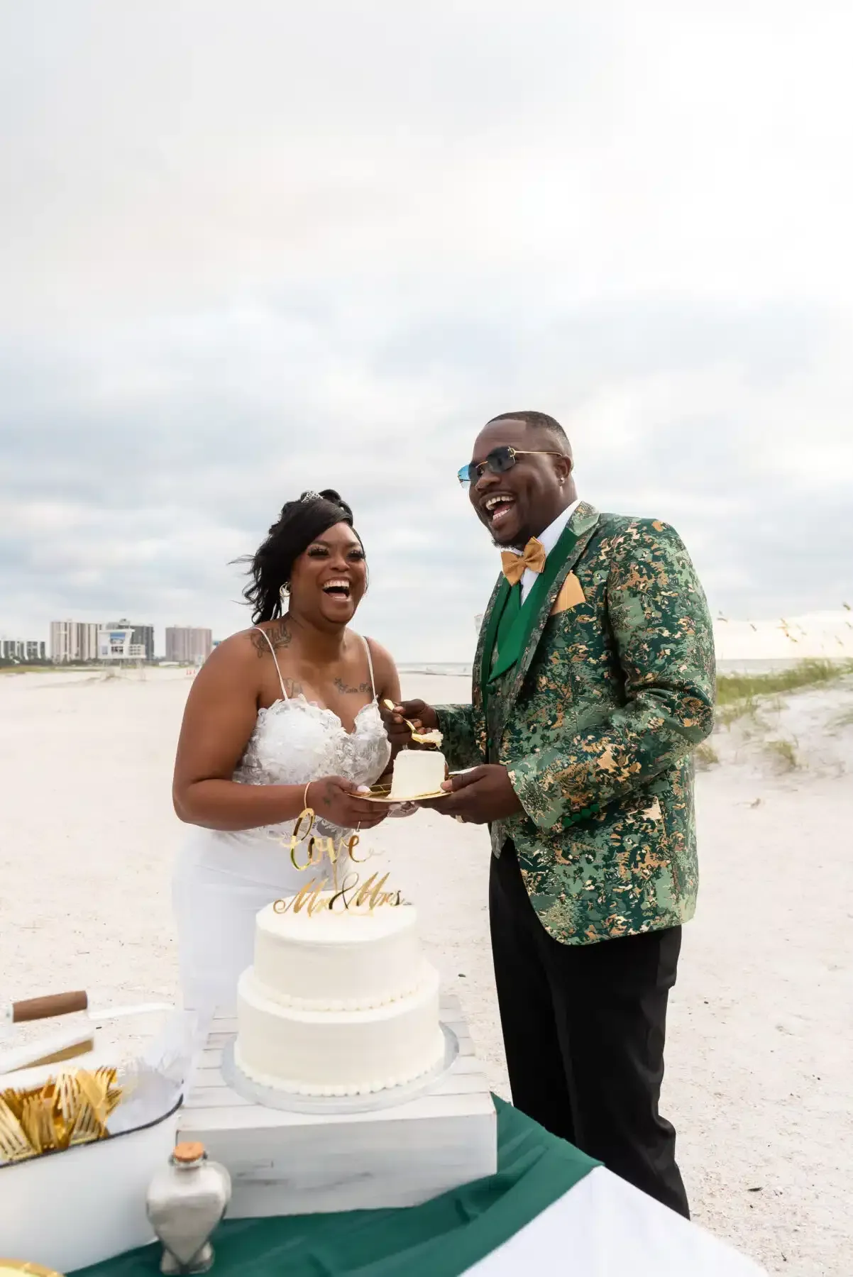 Couple laughing, cutting wedding cake on beach. Groom in green jacket, bride in white dress, cloudy sky.