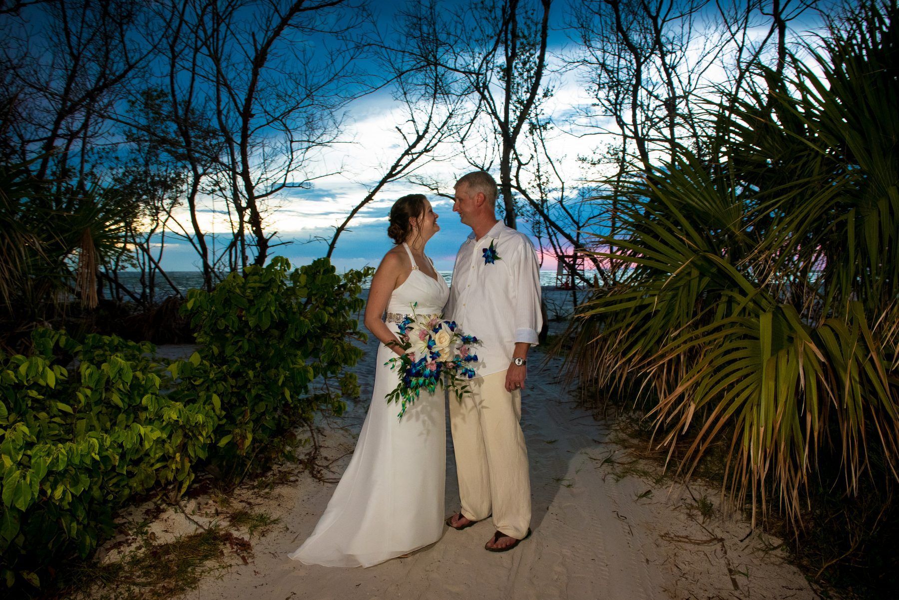 Couple at beach wedding, bride in white dress holding bouquet, groom in linen shirt, path through foliage, dusk.