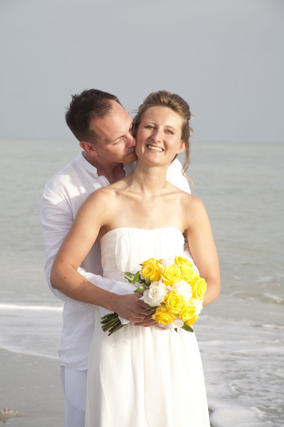 Couple embracing on a beach; woman in white dress holds bouquet; man kisses her cheek.