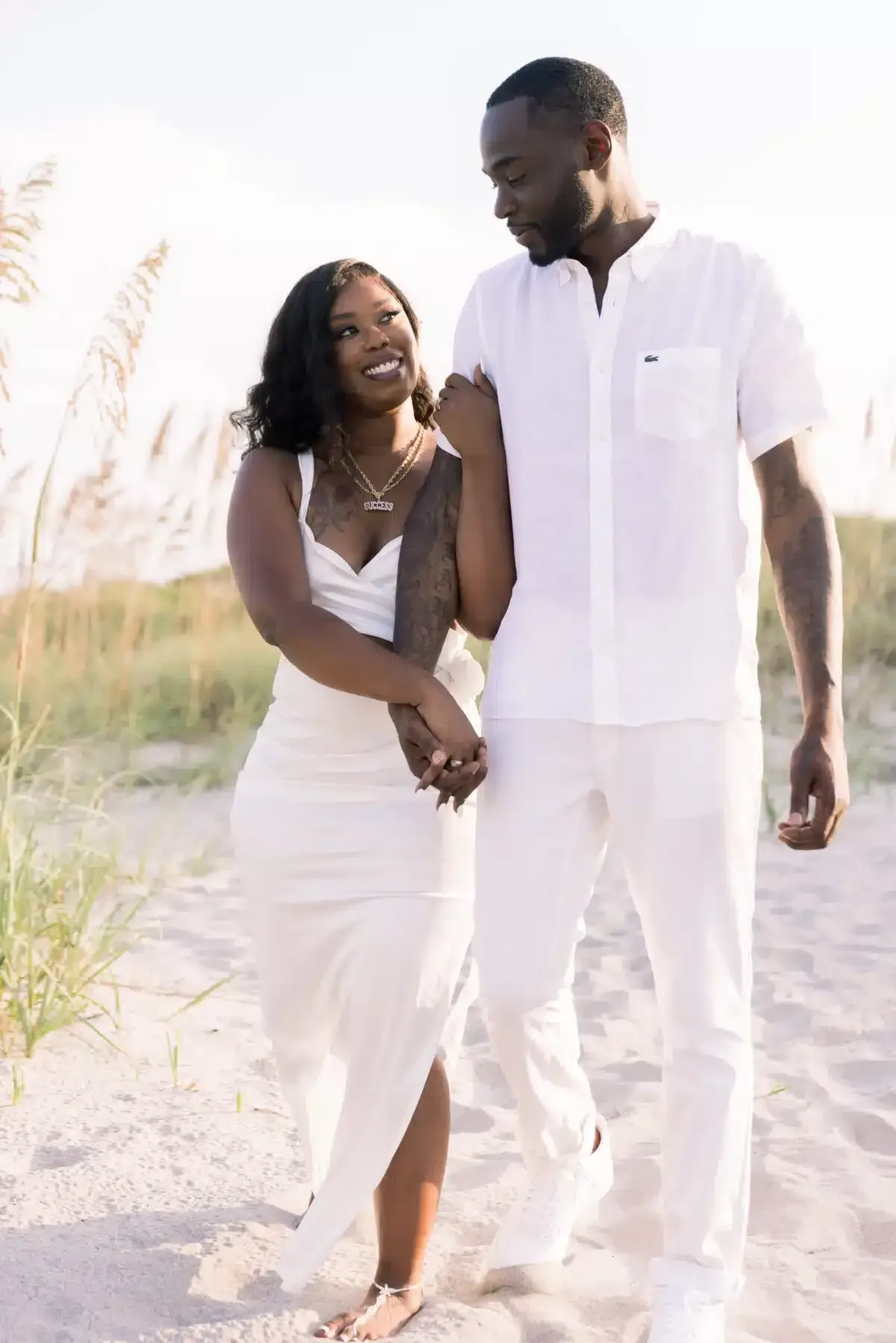 Couple walking on beach, holding hands, smiling at each other. Both wearing white clothing.