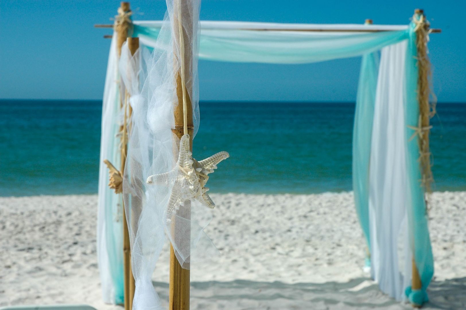 Beach wedding arch decorated with blue and white fabric, starfish, and rope, with ocean background.