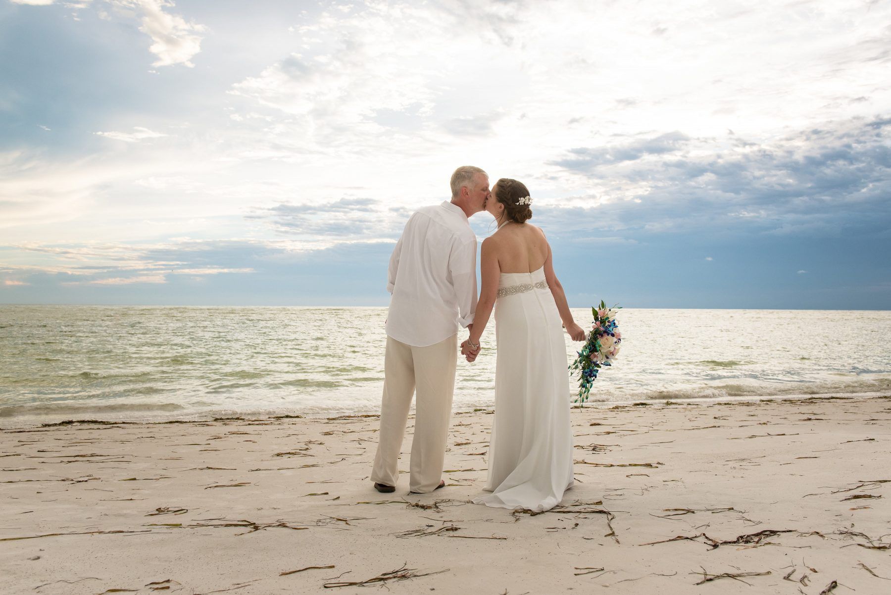 Couple kissing on a beach; woman in wedding dress, man in white shirt and tan pants; ocean and cloudy sky.
