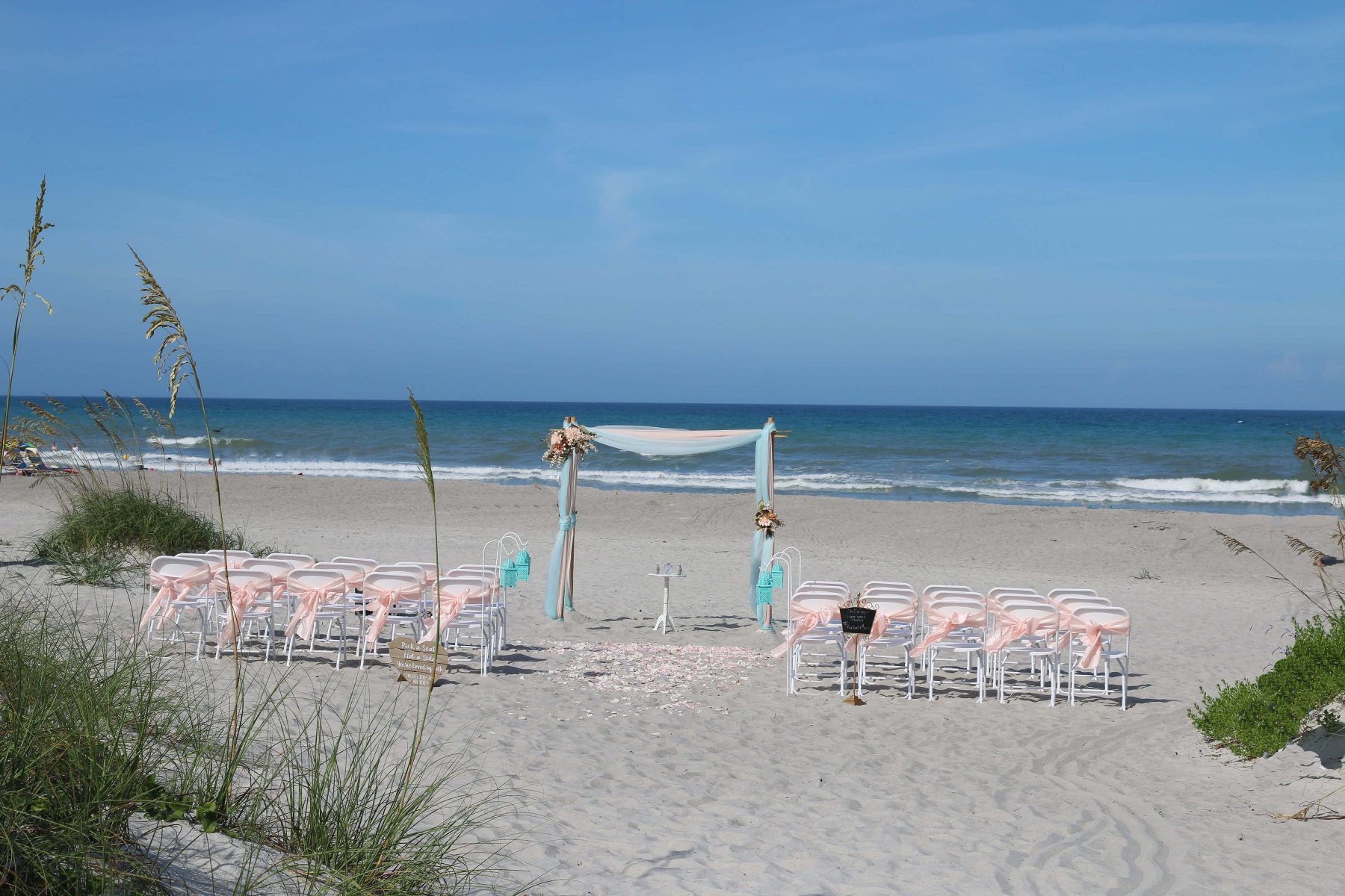 Beach wedding setup with chairs, arch, and ocean view under a blue sky.