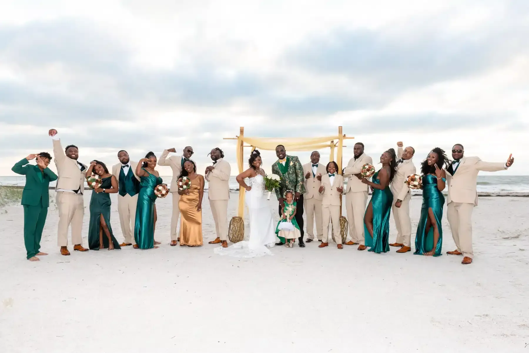 Wedding party on beach; bride in white dress, others in tan and emerald attire, posing near arch.