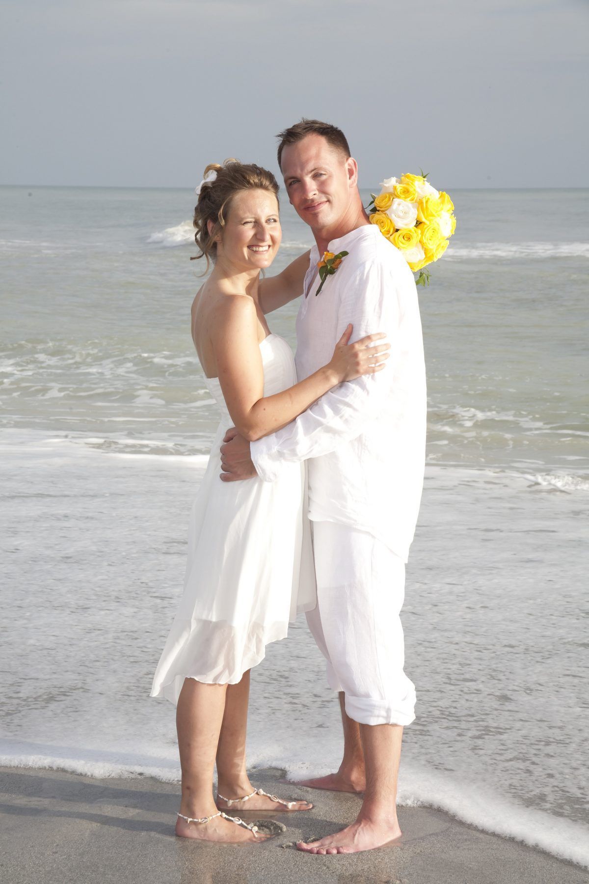 Couple embracing on a beach; the woman wears a white dress, holding a yellow bouquet; the man wears white.