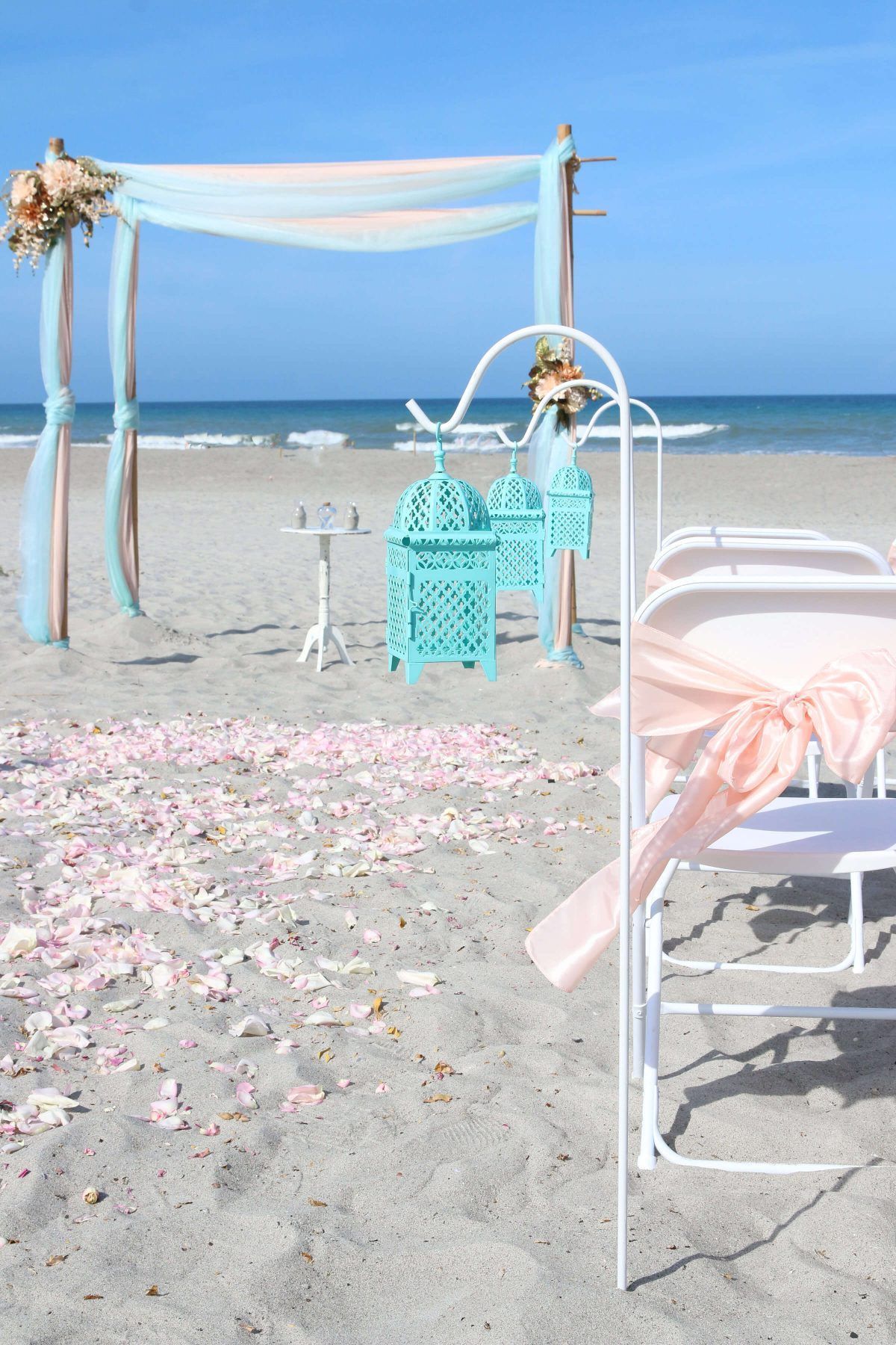Beach wedding setup with arch, chairs, and lanterns. Pink and blue decor on the sand.