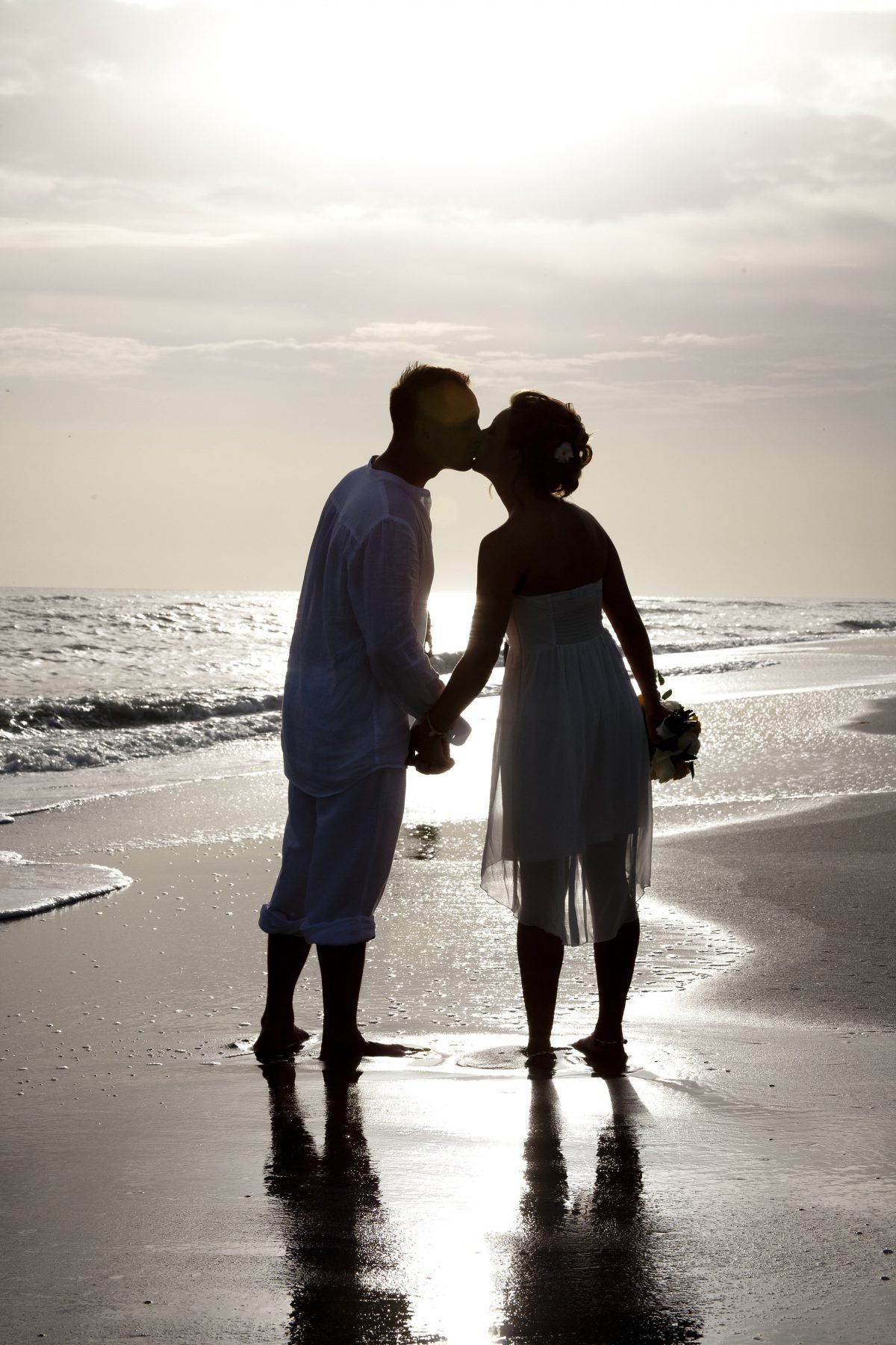 Couple kissing on a beach at sunset, silhouetted against the sky. Holding hands, with water and reflections.