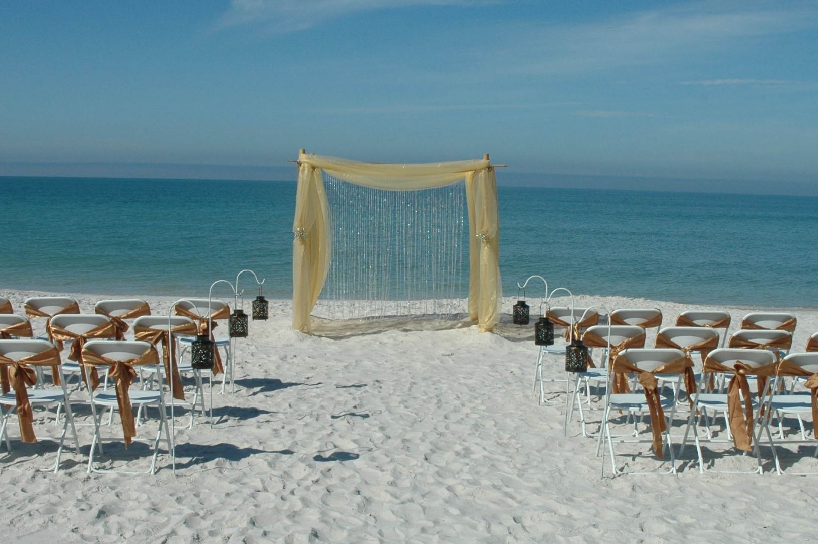 Wedding ceremony setup on a white sandy beach with ocean backdrop; chairs, arch, lanterns.