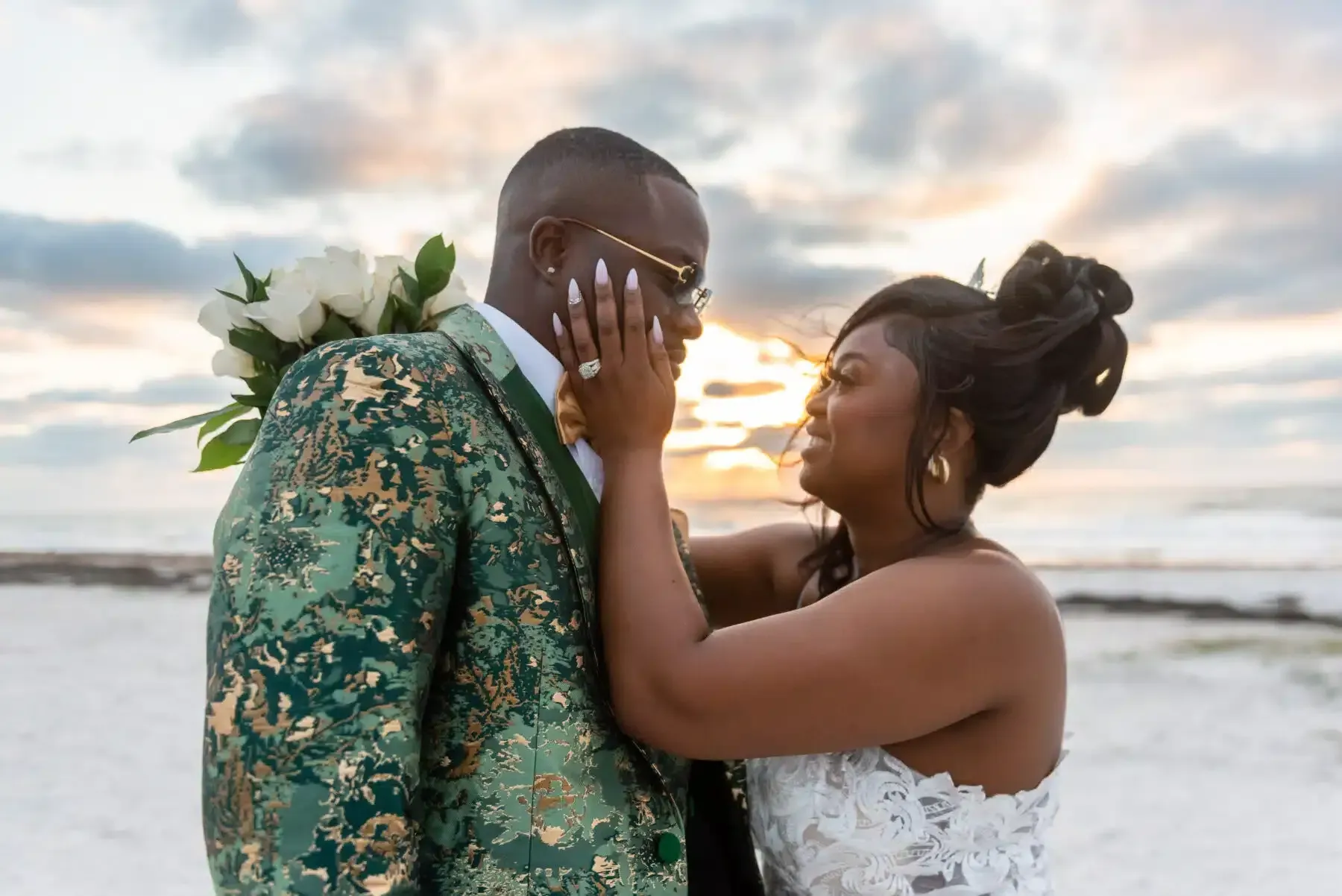 Bride touching groom's face on beach at sunset. Groom in green suit, holding flowers.