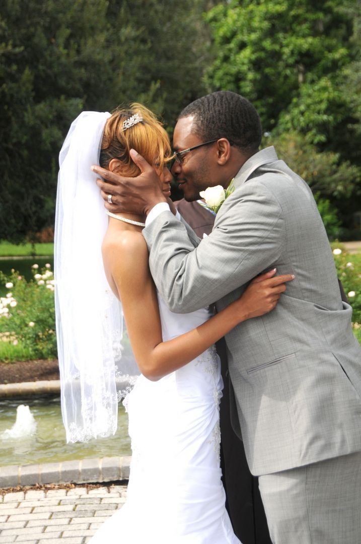 Bride and groom embracing, about to kiss. Bride in white dress and veil, groom in gray suit, outdoors near a fountain.