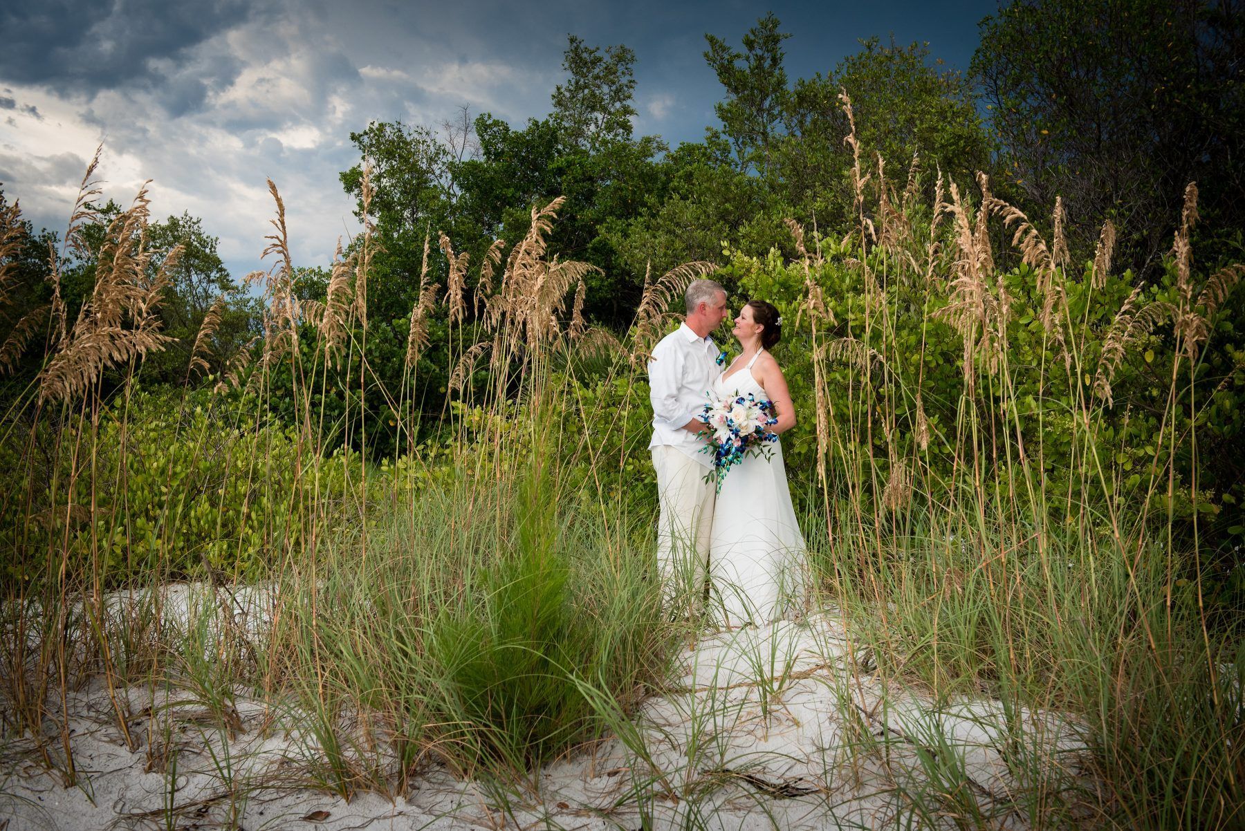 Bride and groom on a beach, facing each other. Surrounded by sea oats and green foliage under a cloudy sky.