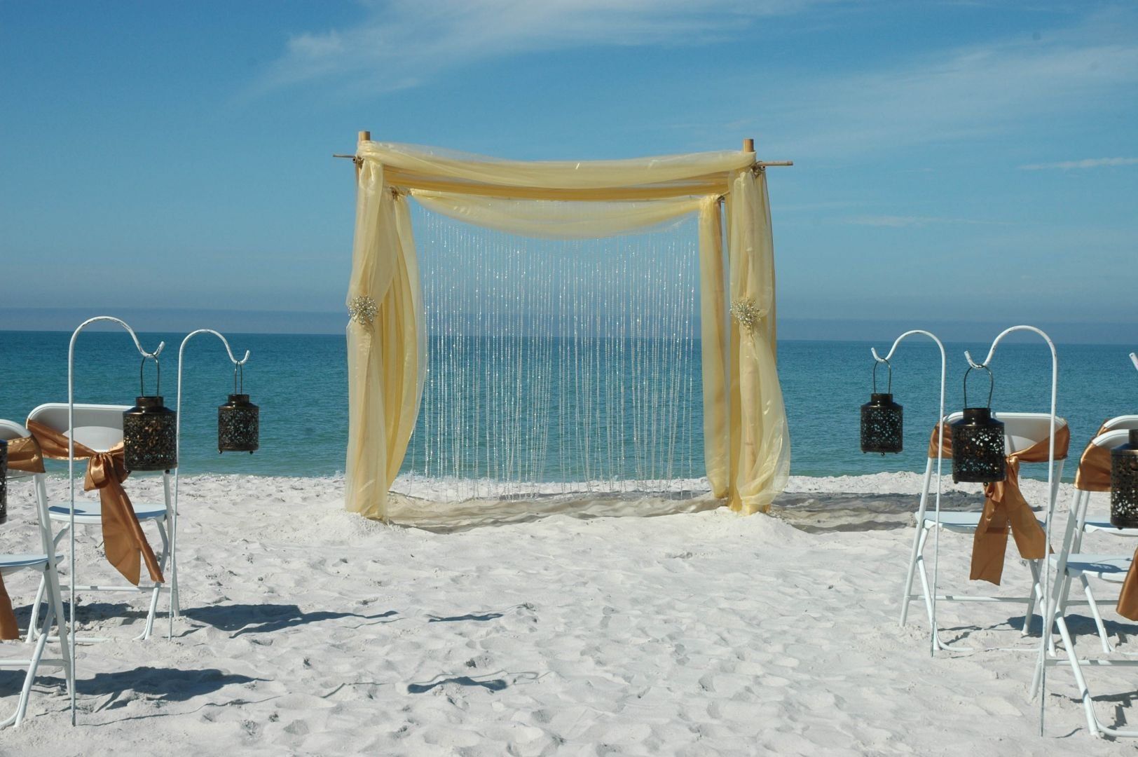 Beach wedding setup with draped arch, white chairs, lanterns, ocean backdrop.