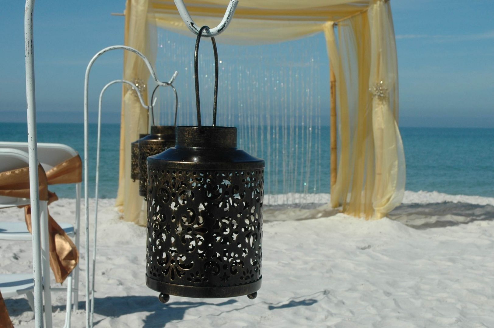 Beach wedding setting with ornate lanterns, sheer fabric, and ocean backdrop.