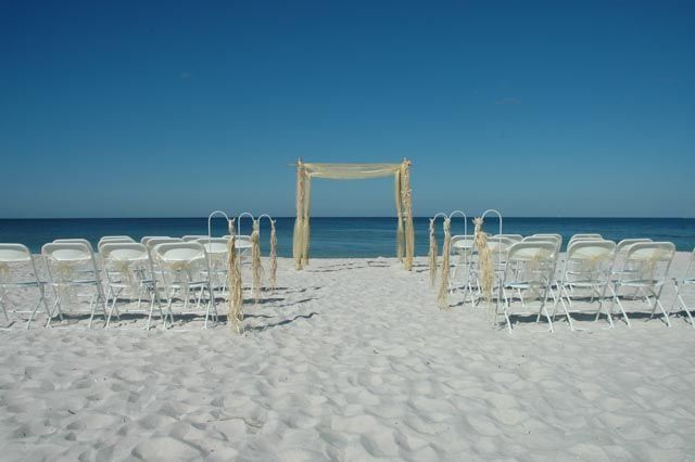 Wedding ceremony setup on a sandy beach with white chairs facing a decorated arch, ocean, and clear blue sky.