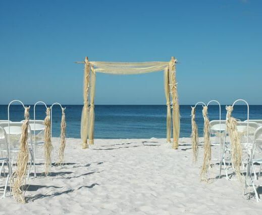 Wedding ceremony setup on white sand beach, arch, chairs, ocean backdrop.