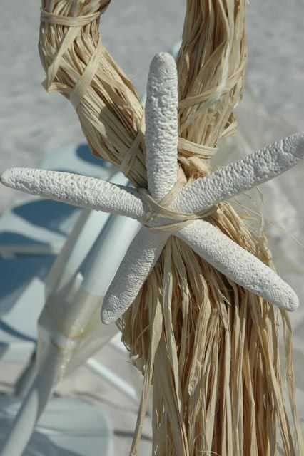 White starfish tied to raffia-covered arch, beach setting with white chair in background.