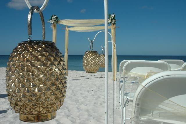 Beach wedding setup with gold lanterns, white chairs, and arch against blue sky and ocean.