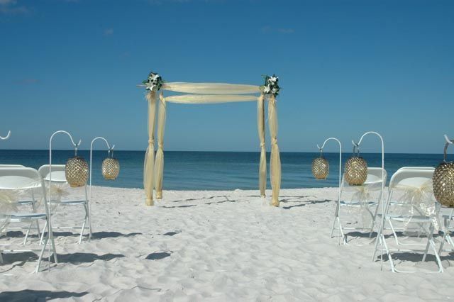 Beach wedding setup with arch, chairs, and lanterns. Blue sky and ocean in background.