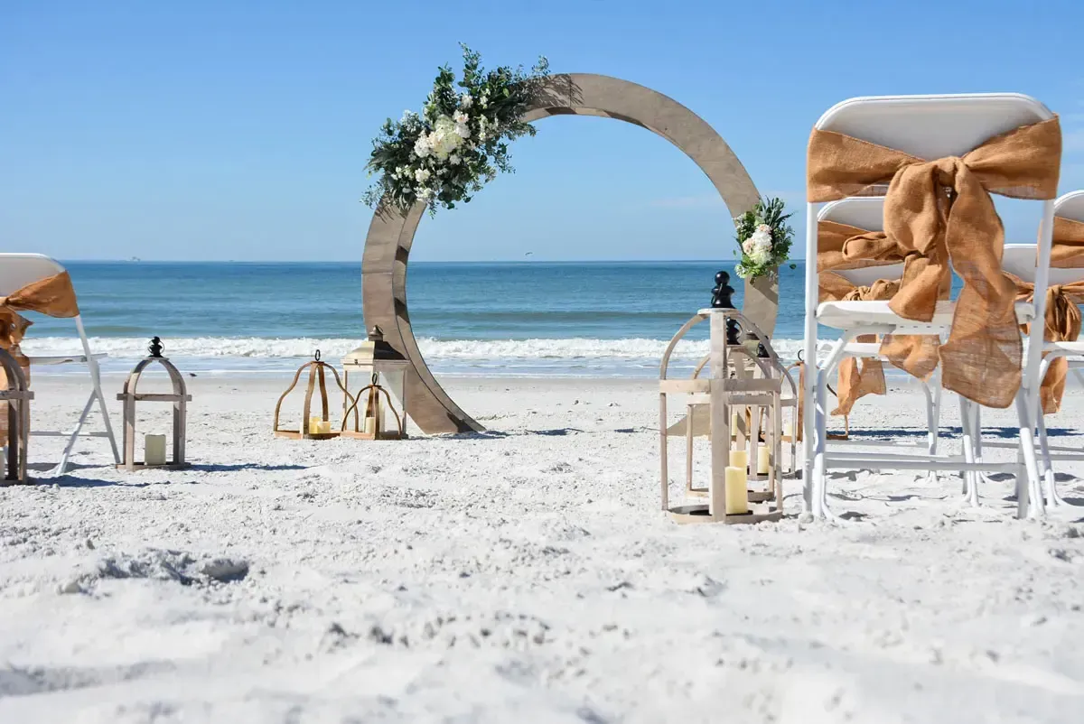 Beach wedding setup: circular arch decorated with flowers, chairs with burlap bows, lanterns, and ocean backdrop.