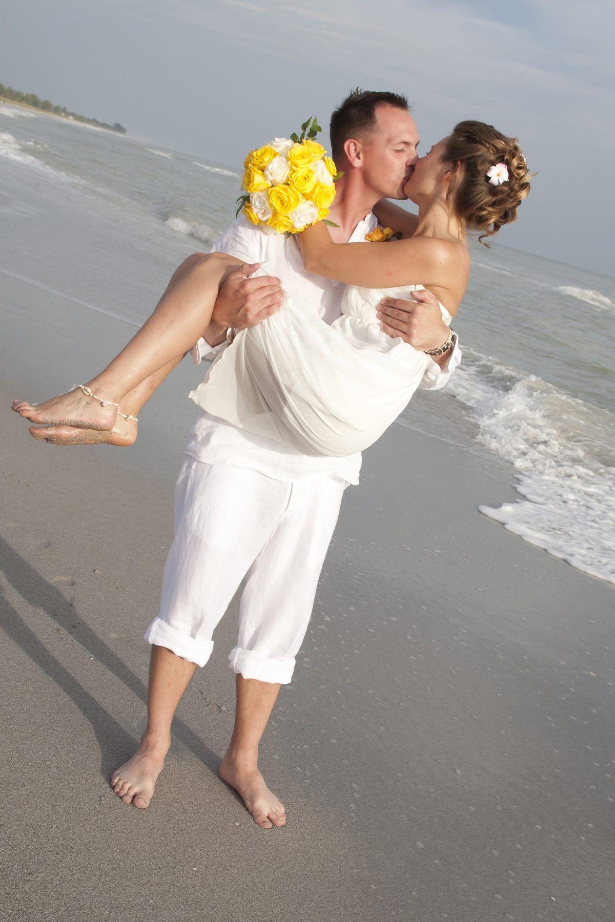 Groom carries bride, kissing, on beach. Bride holds yellow bouquet. Both wear white, ocean background.
