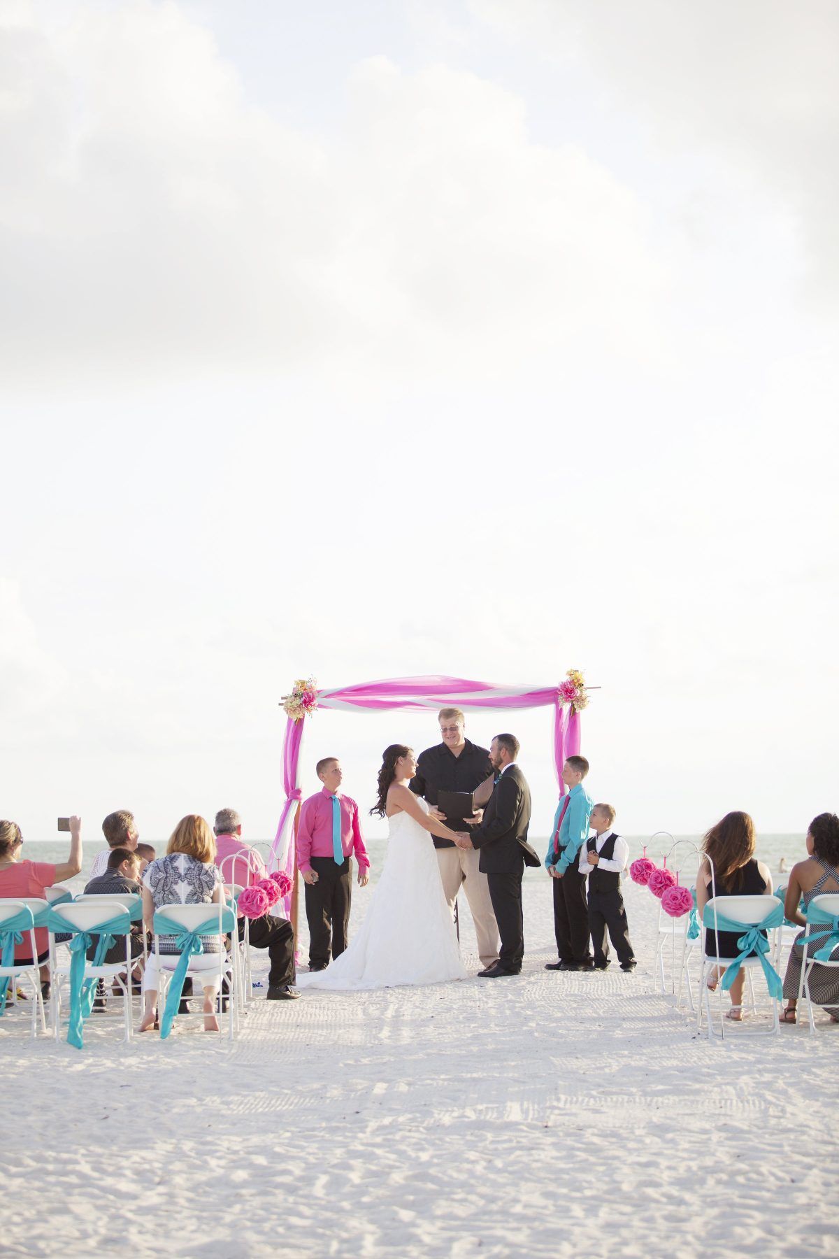 Beach wedding ceremony with couple, officiant, and guests under a pink arch.