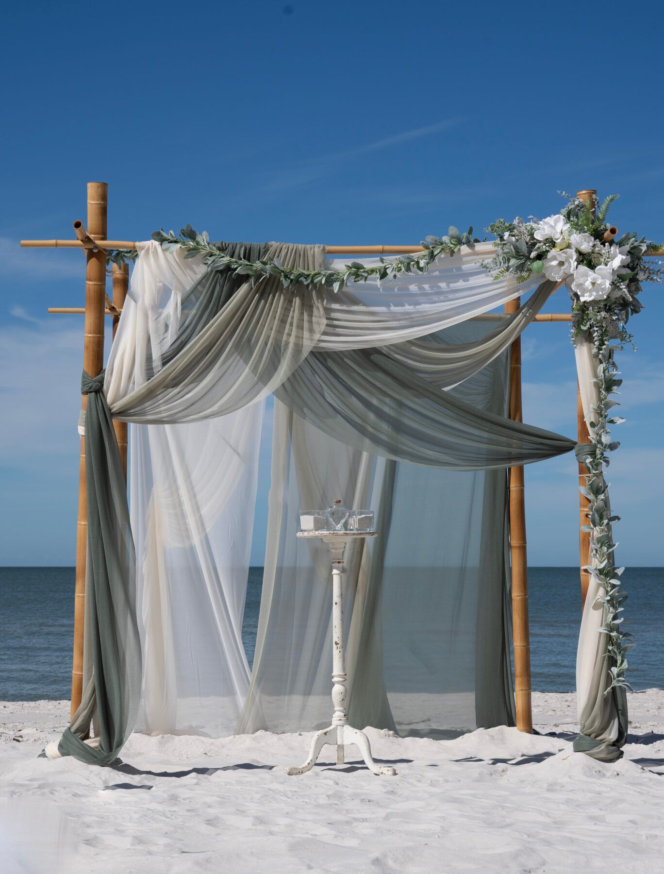 Beach wedding arbor with draped fabric and floral accents on a sunny day.