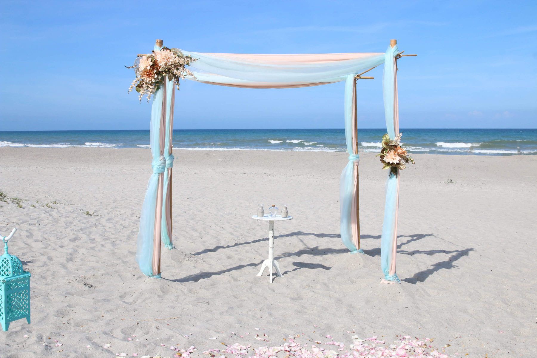 Beach wedding setup with draped fabric, floral accents, and a stand, with the ocean in the background.