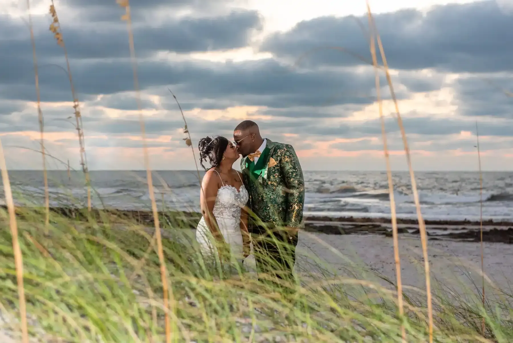 Couple kisses on a beach at sunset, behind tall grass.