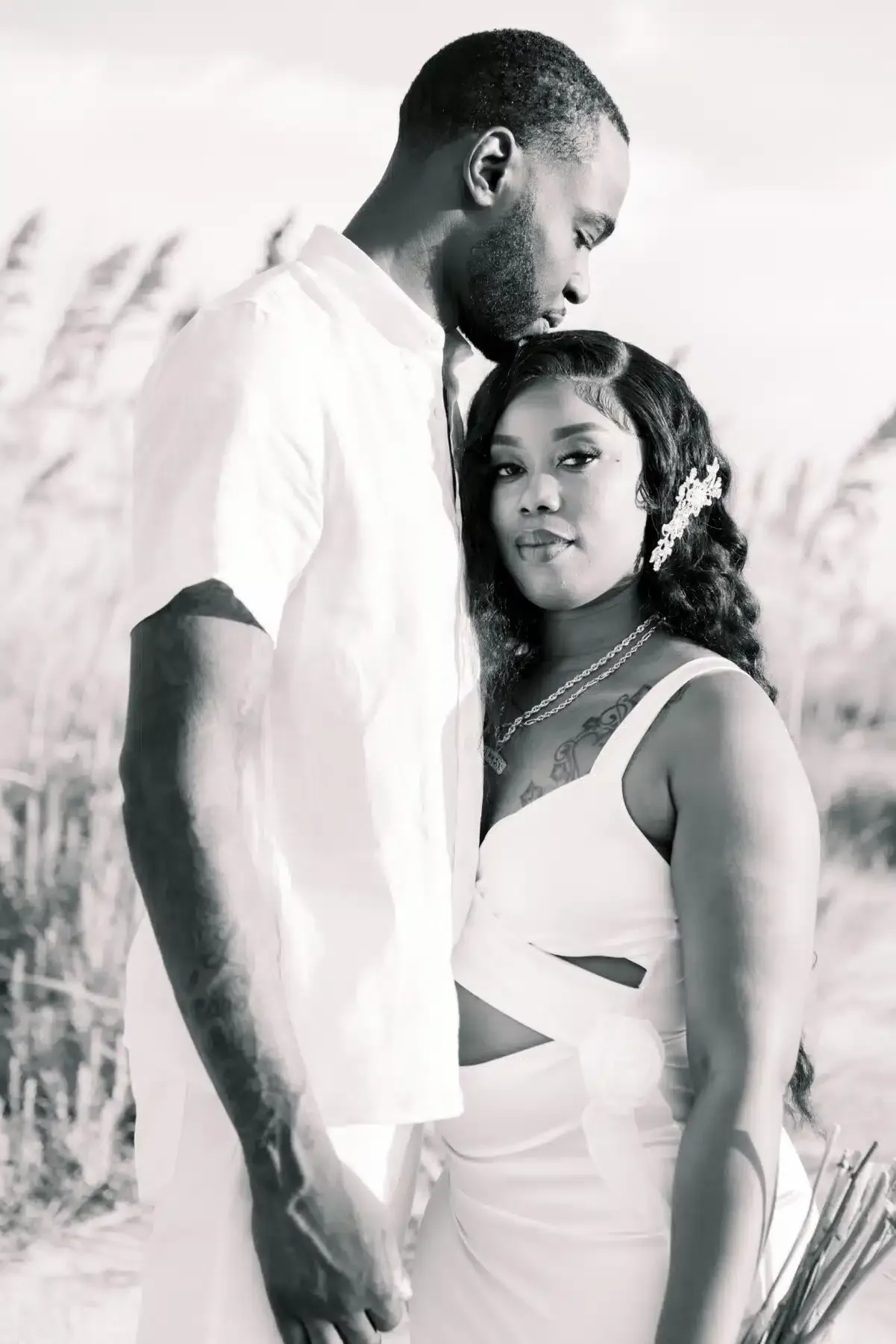 Couple embracing on a beach, woman in white dress, man in white shirt, tall grass background.