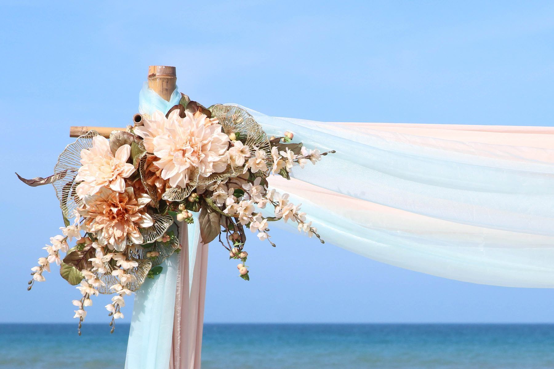 Wedding arch with peach and blue fabric, adorned with flowers, on beach.