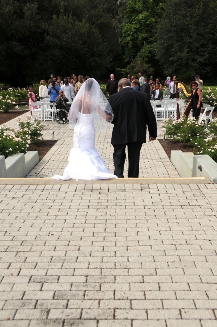 Bride in white dress walks down stone steps, held by person in black suit, toward a wedding ceremony.